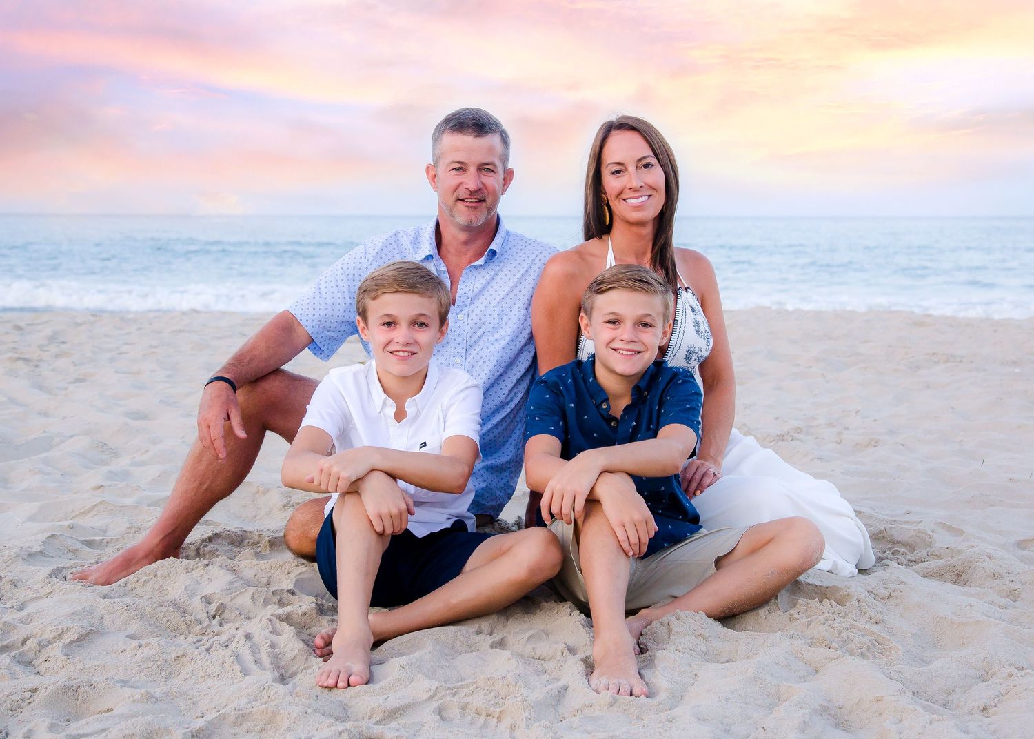 Happy family of four posing on sandy beach at sunset with two boys in matching outfits.