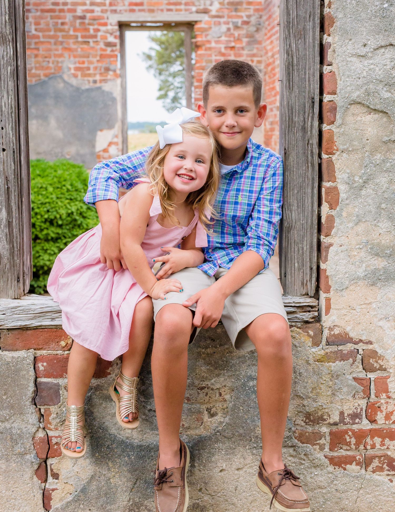 Young boy and girl siblings posing together in a rustic brick doorway in coordinating outfits.