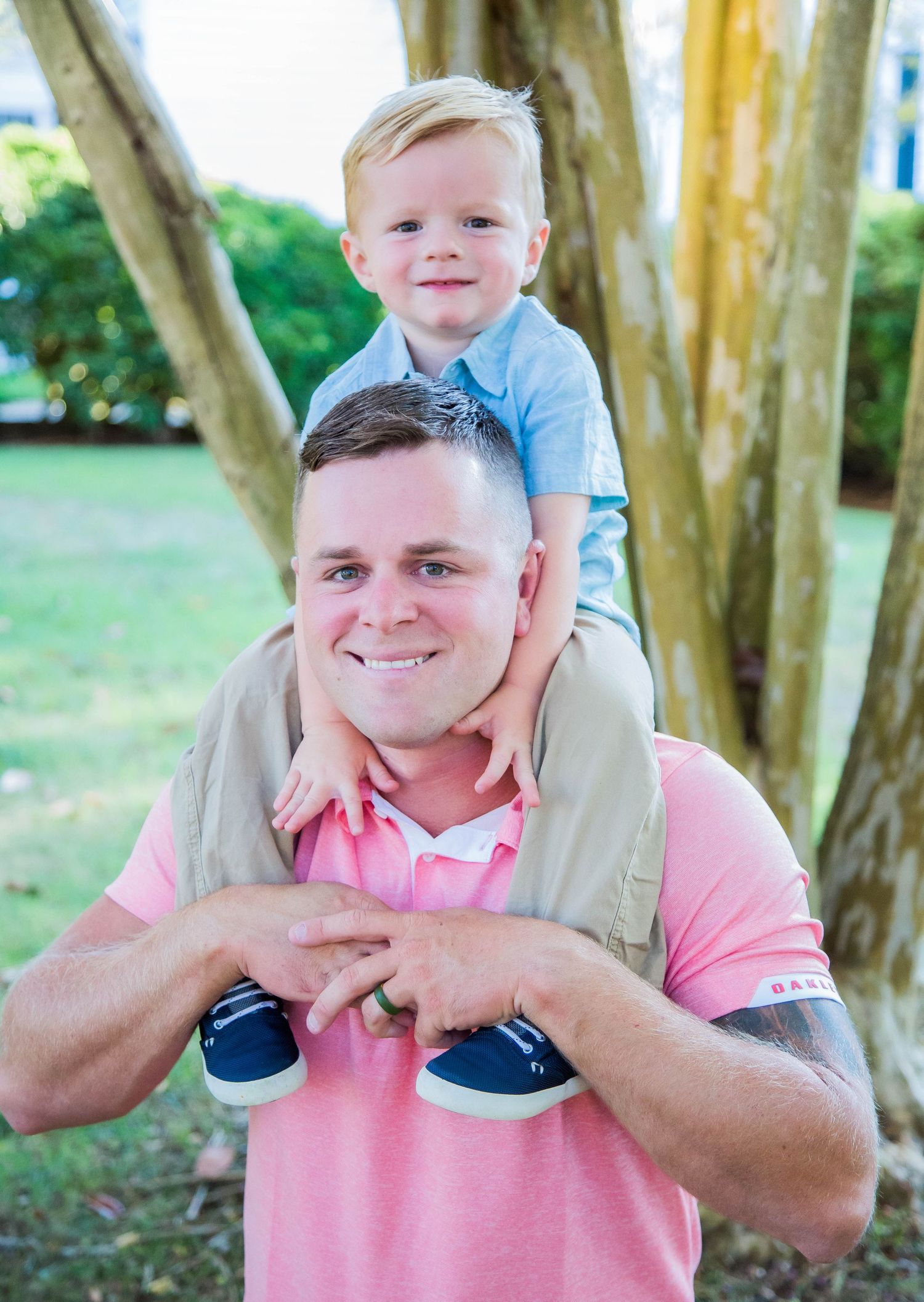 Father in pink polo shirt carrying smiling toddler boy on his shoulders outdoors.