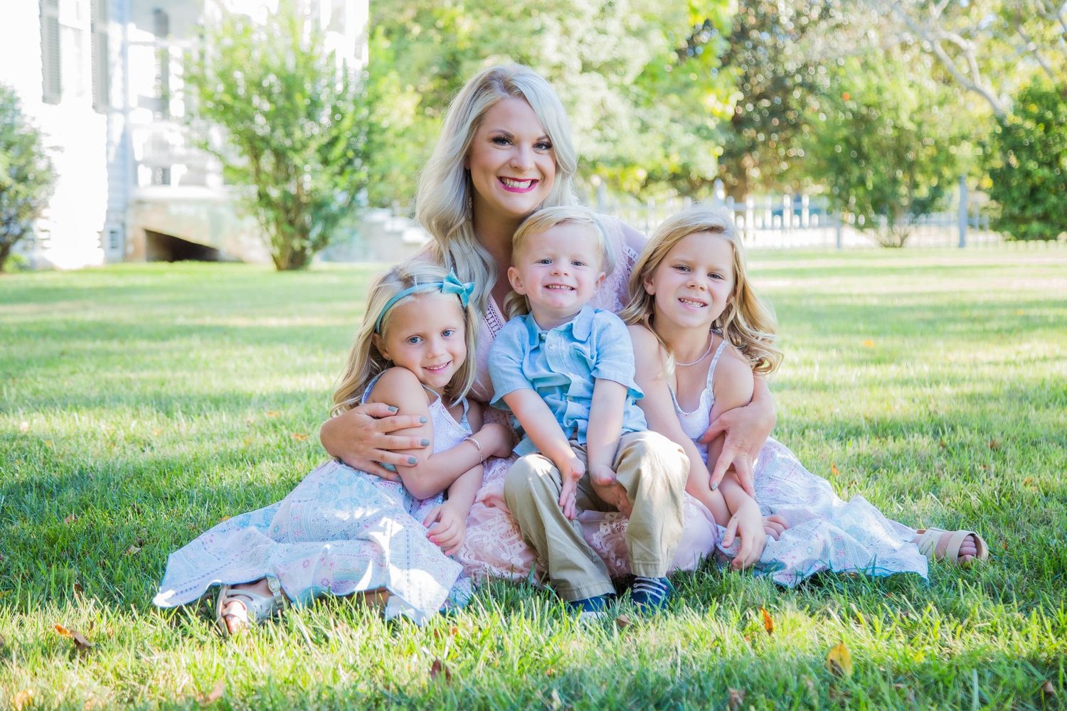 Smiling blonde mother sitting on grass with three young children in coordinating outfits for a family portrait.
