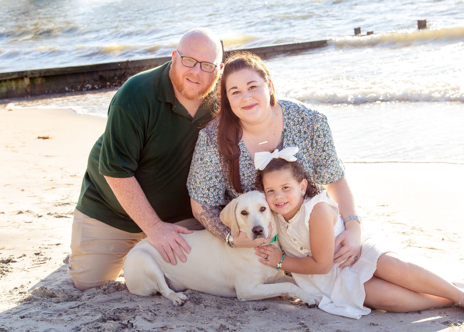 Smiling family with a yellow Labrador posing together on a sandy beach at sunset.