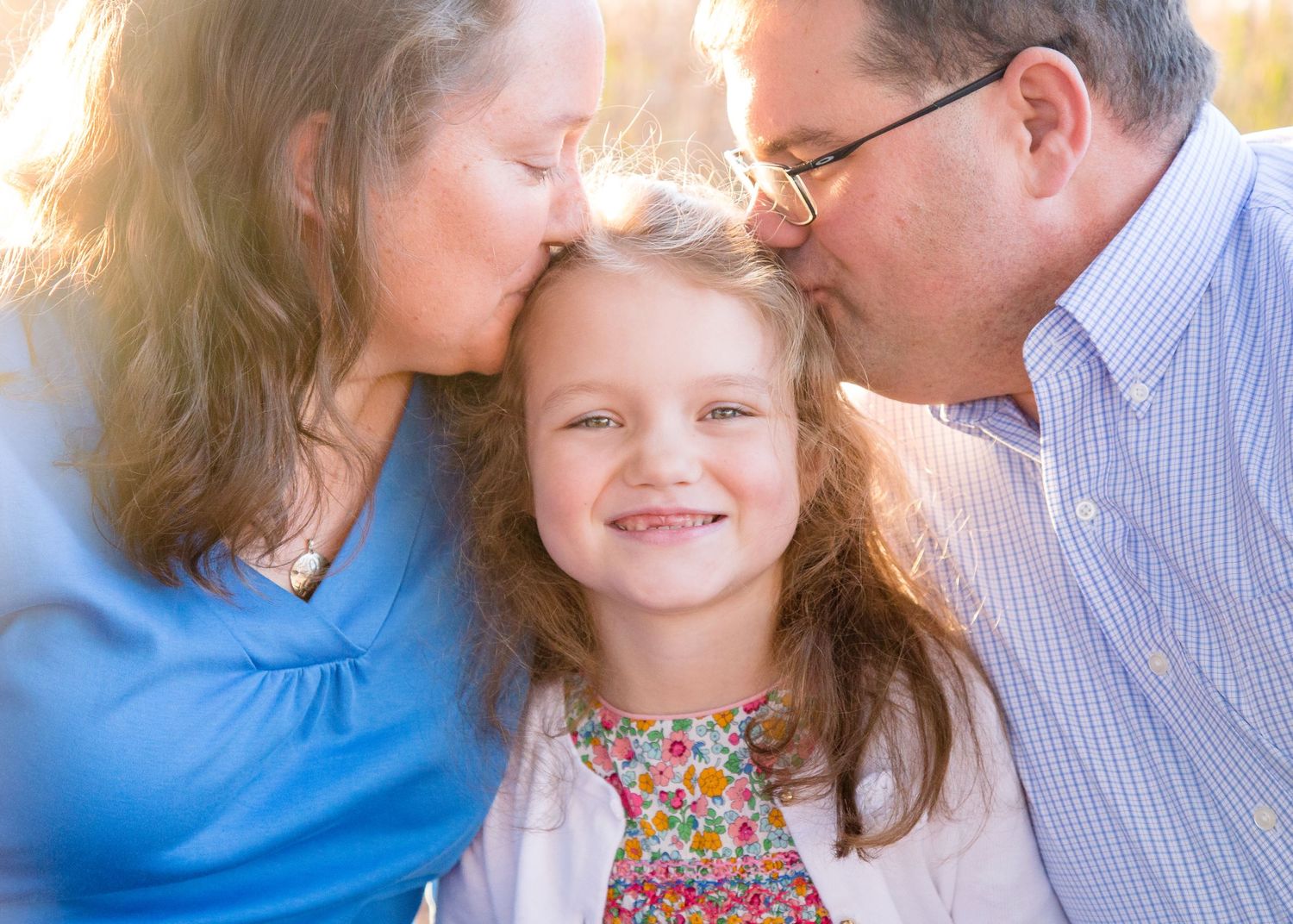 Smiling young girl receiving kisses on the cheeks from both parents outdoors.