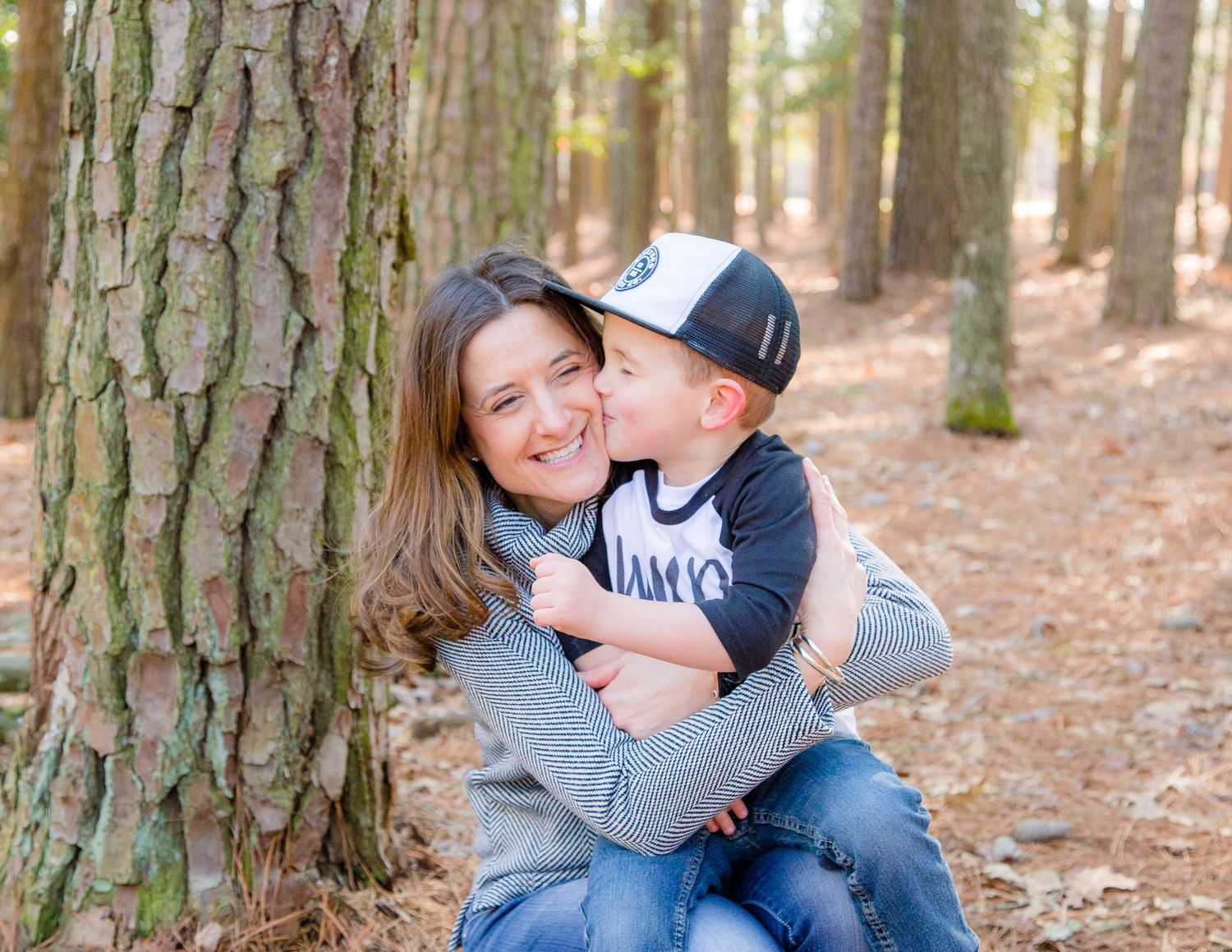 Loving mother hugging her young son beside a pine tree in a sunlit forest.