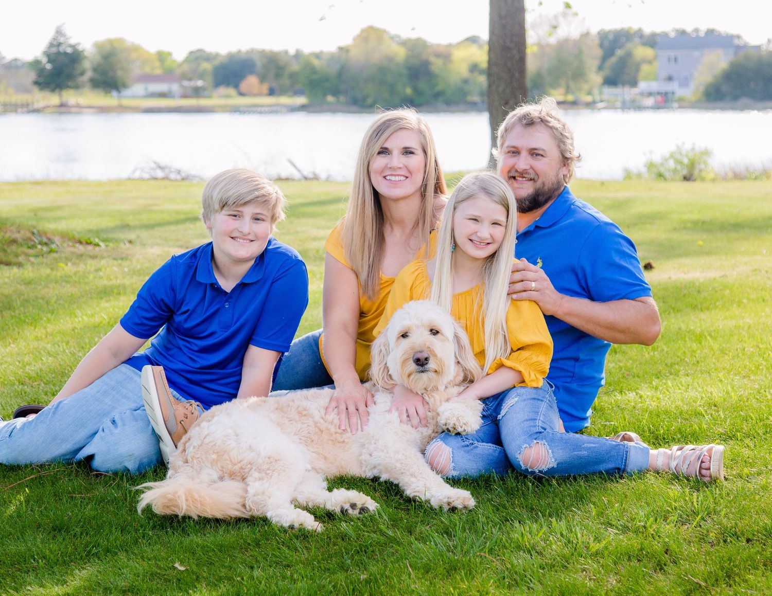 Happy family of four with a fluffy dog sitting on green grass near a lake.