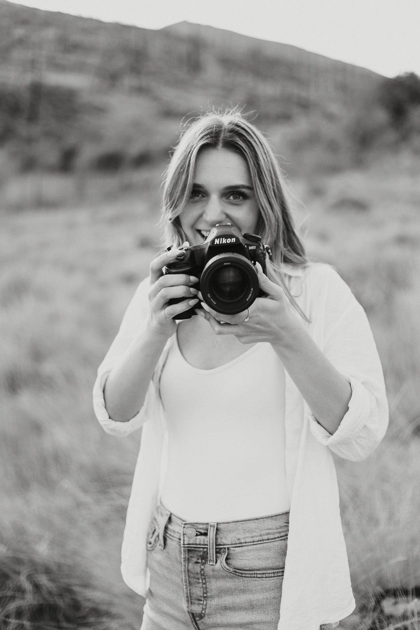 A photographer in casual white attire holds up a professional camera in black and white.