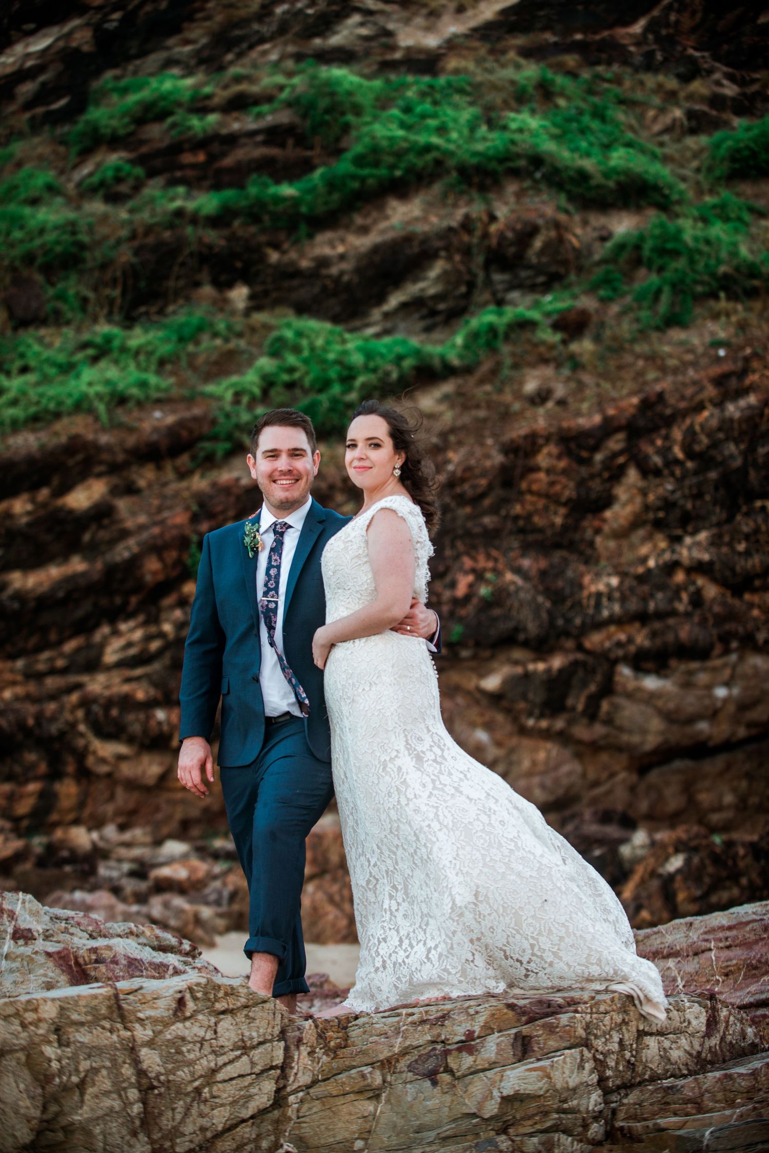 A wedding couple posing on rocky terrain with green foliage in the background.
