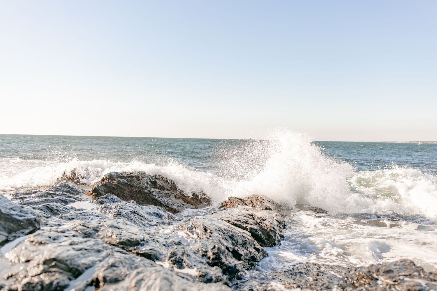 Powerful ocean wave splashing over coastal rocks under a bright clear sky.