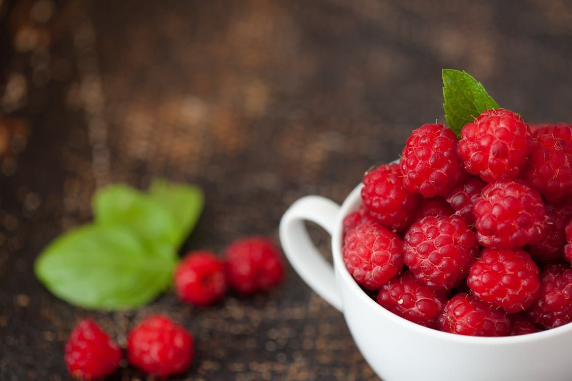 Fresh red raspberries piled in white ceramic cup with green leaves.