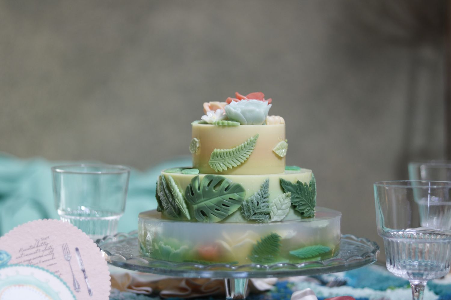 Three-tiered tropical-themed wedding cake with green leaf details on a glass cake stand.