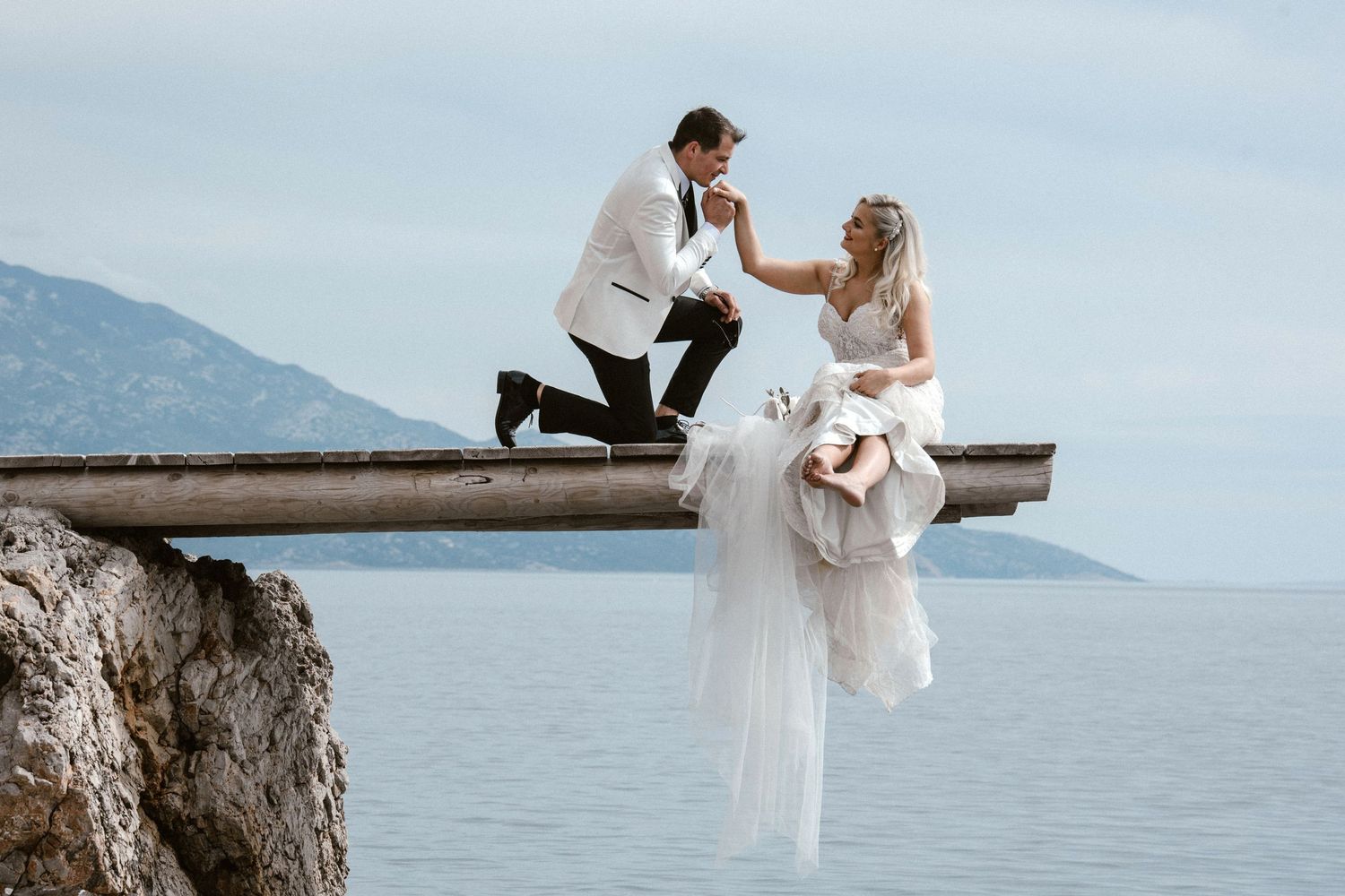 Adventurous wedding photo on cliff edge overlooking ocean and mountains.