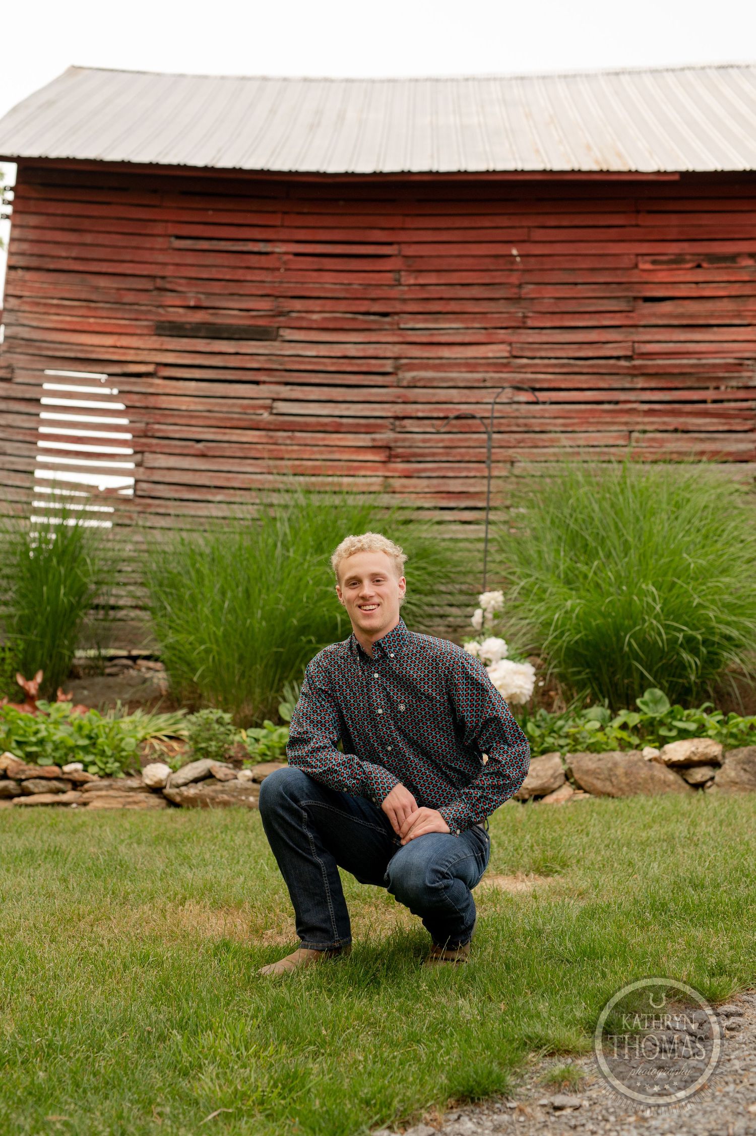 Standing pose in front of rustic red barn wall with tall ornamental grasses and rocks in foreground.
