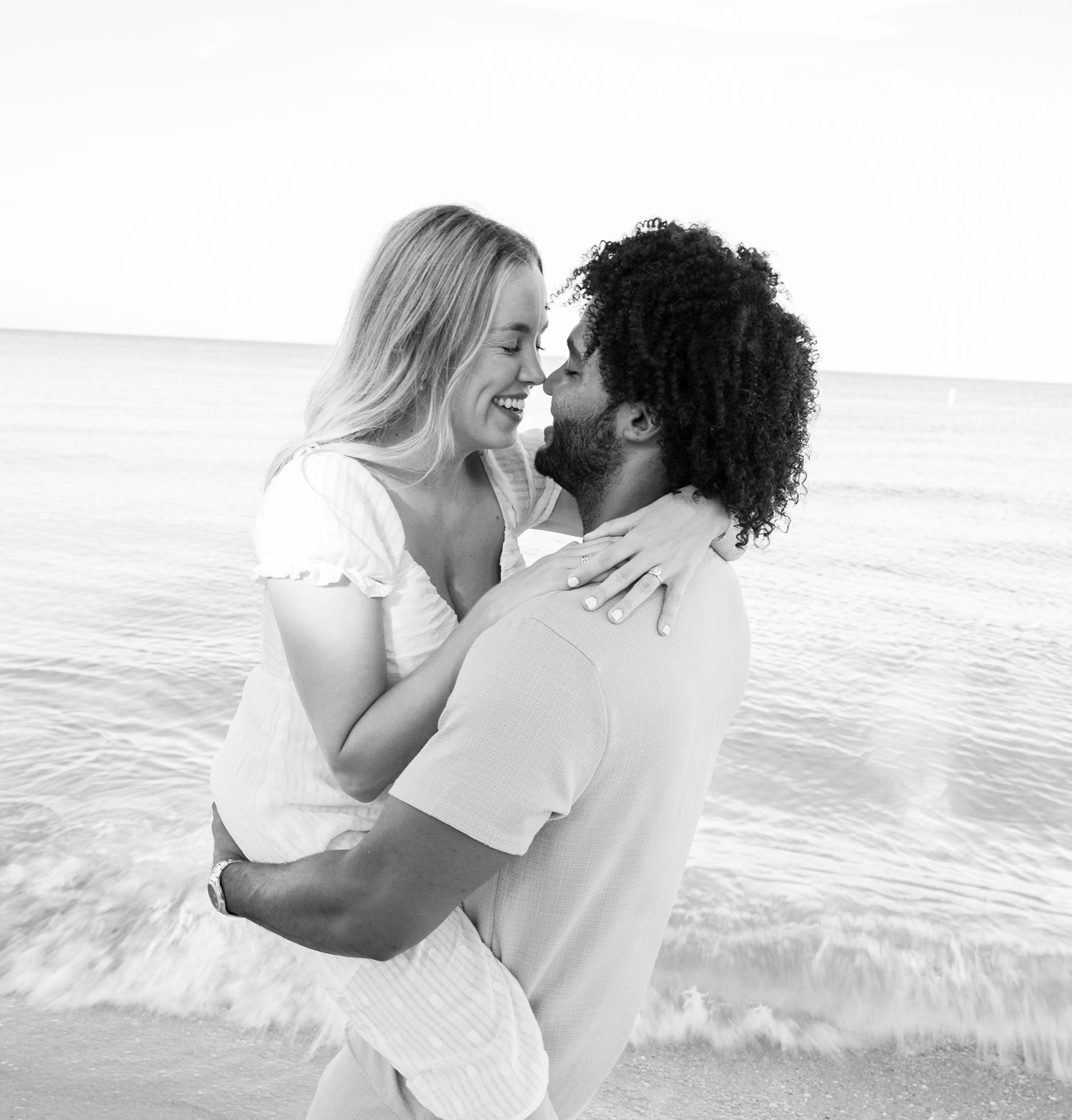 A romantic black and white beach portrait of a couple embracing and laughing by the ocean.