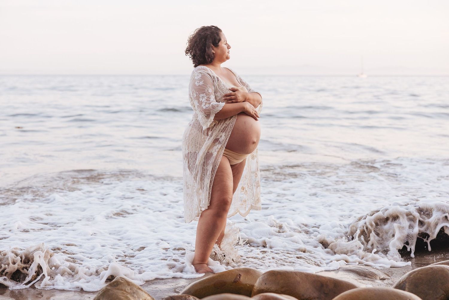 Ethereal maternity photo of an expectant mother in a lace dress standing on rocky beach at sunset.