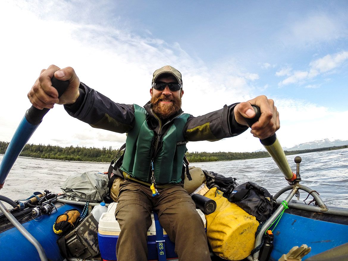Smiling Arkansas nature photographer, Alex Kent, facing the camera rowing a blue raft with his hand holding the oars.