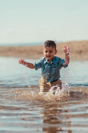 A child splashes joyfully in shallow beach water wearing a blue shirt and khaki shorts.