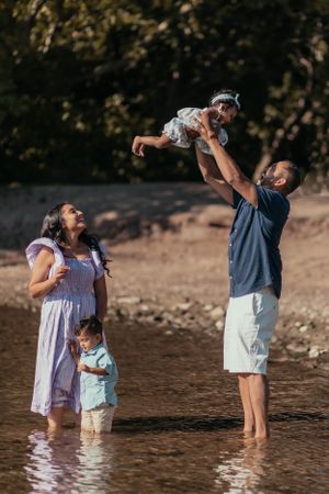 Joyful family playing in shallow water at beach with parent lifting child high in air.