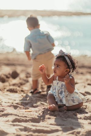 Small child in floral outfit sitting on sandy beach with ocean waves behind.