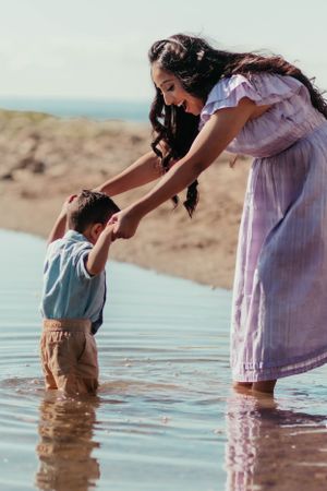 A woman in a lavender dress helps a small child stand in shallow beach water.