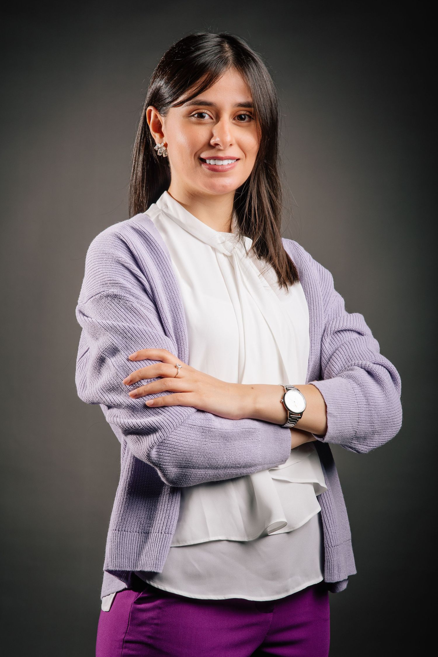 Professional studio portrait showing a person in a lavender cardigan over white blouse with purple pants, posing confidently.
