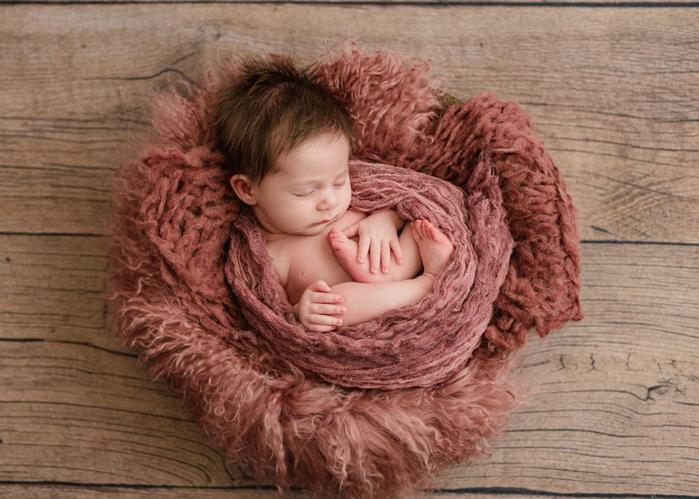 Sleeping newborn wrapped in pink swaddle on wooden surface with fluffy mauve blanket.