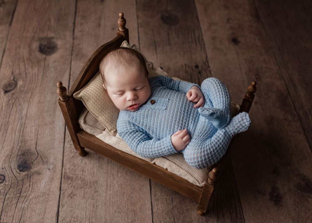 Sleeping baby wearing blue knit outfit lying in miniature wooden bed.