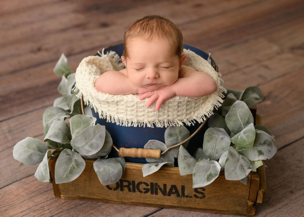 Sleeping newborn in rustic wooden crate with eucalyptus leaves and cream blanket.