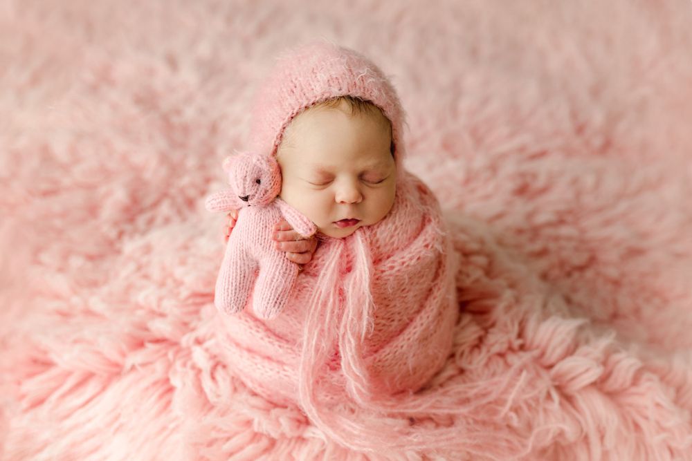 Peaceful newborn baby wearing pink knit outfit sleeping on fluffy pink backdrop.