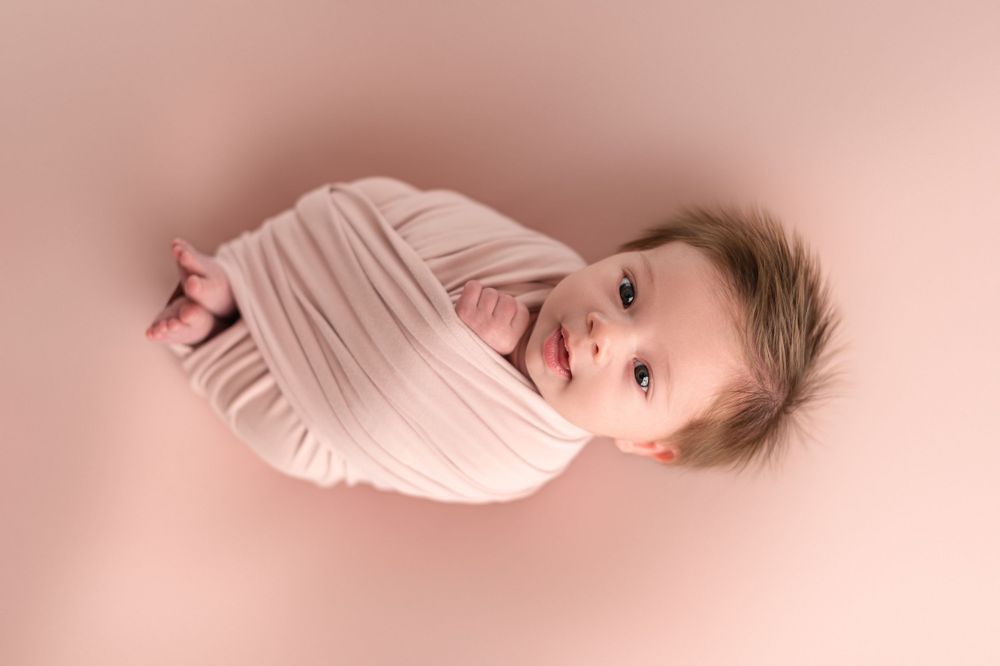 Newborn wrapped in soft pink fabric against rose colored background.