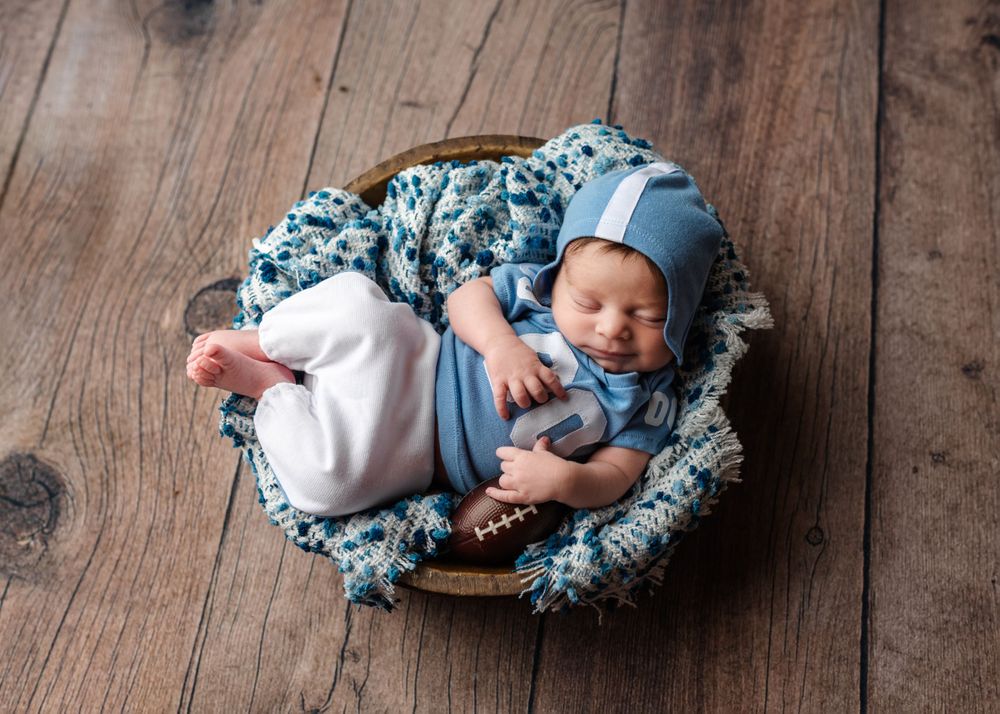 Sleeping newborn in blue outfit lying on floral blanket in rustic wooden basket.