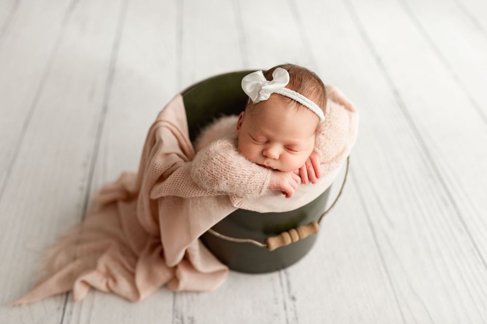 Sleeping newborn wrapped in pink blanket resting in olive green bowl on white wooden floor.
