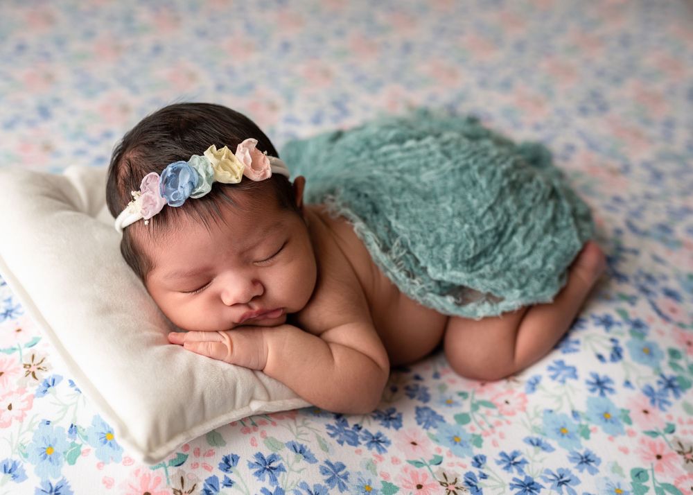 Sleeping newborn wearing floral headband and teal wrap on floral background.