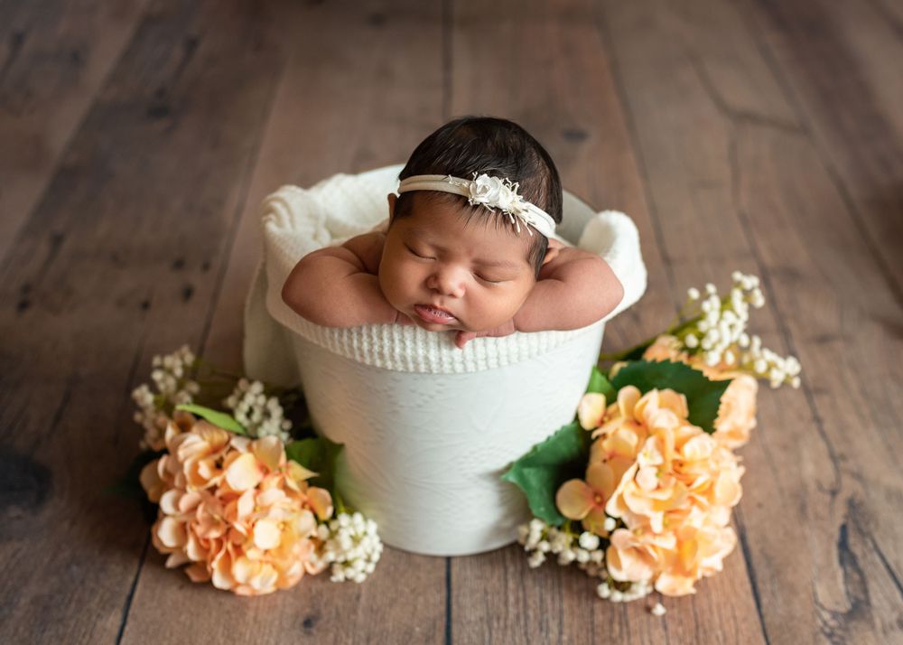 Sleeping newborn in white bucket with floral decorations on wooden floor.