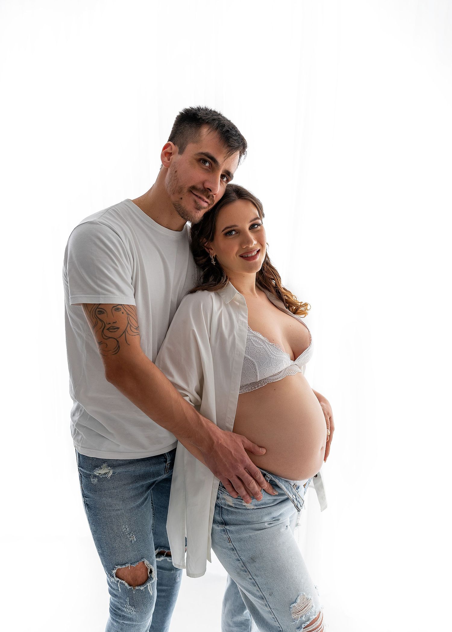 Expecting couple in white shirts and jeans pose together against white background.