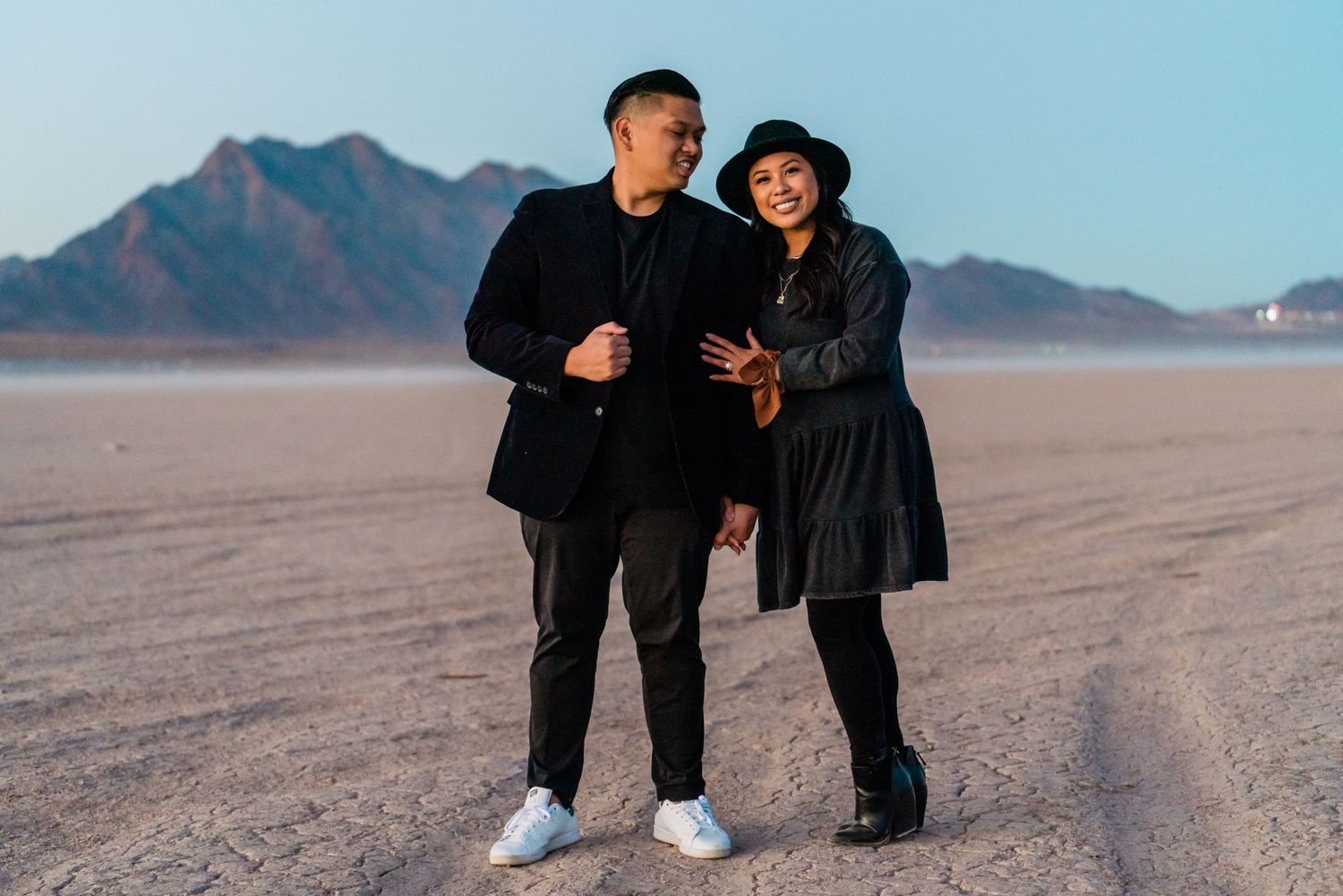 A couple poses together in stylish black outfits against a desert landscape at sunset.