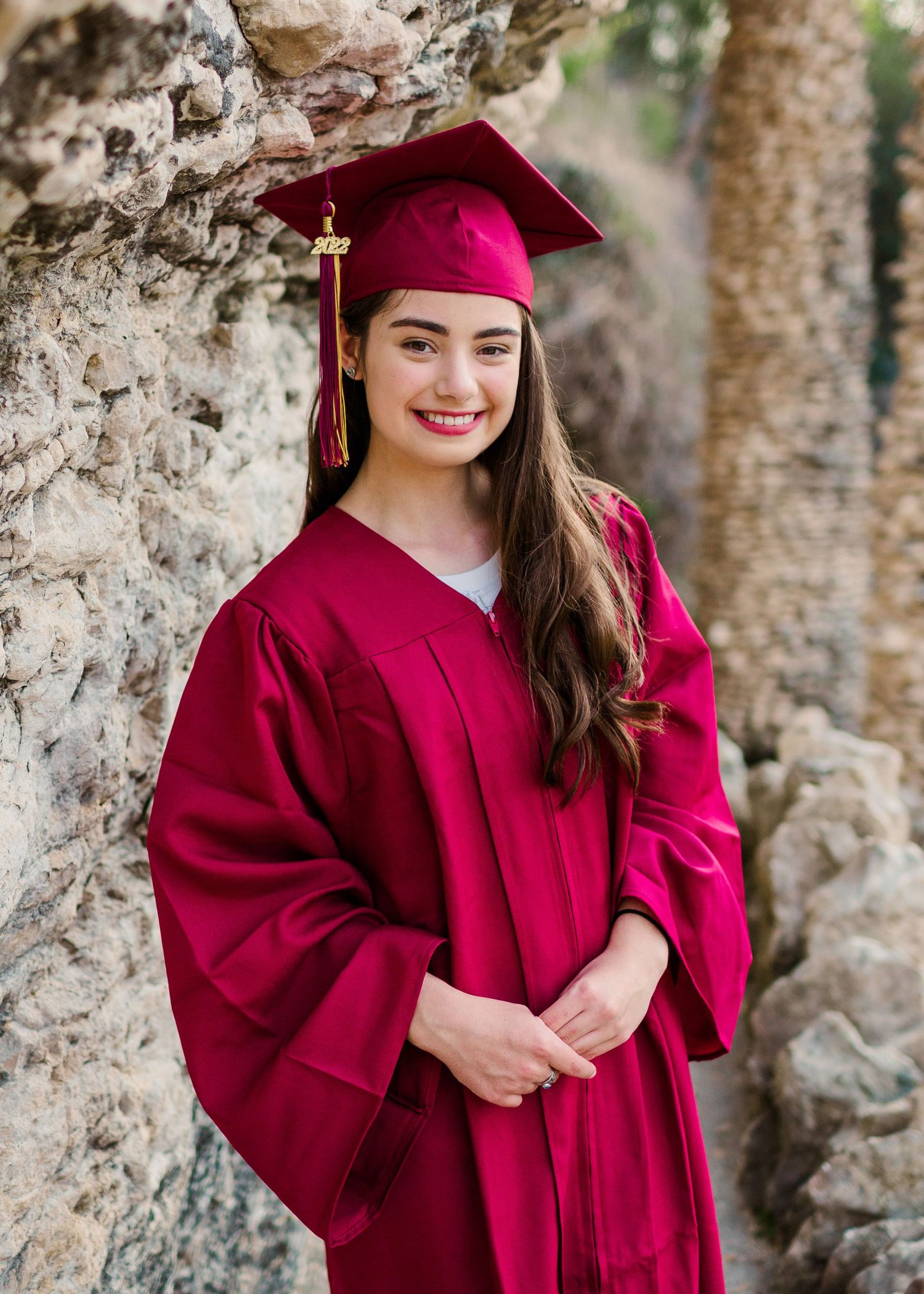 Graduate wearing burgundy cap and gown poses against stone wall during graduation photoshoot.