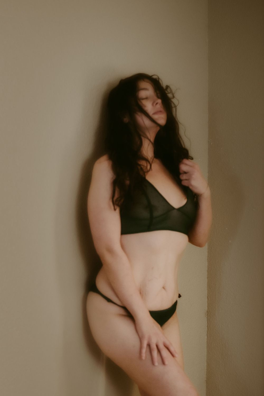 Artistic boudoir photo of a figure in black lingerie posed against a beige wall in soft lighting.