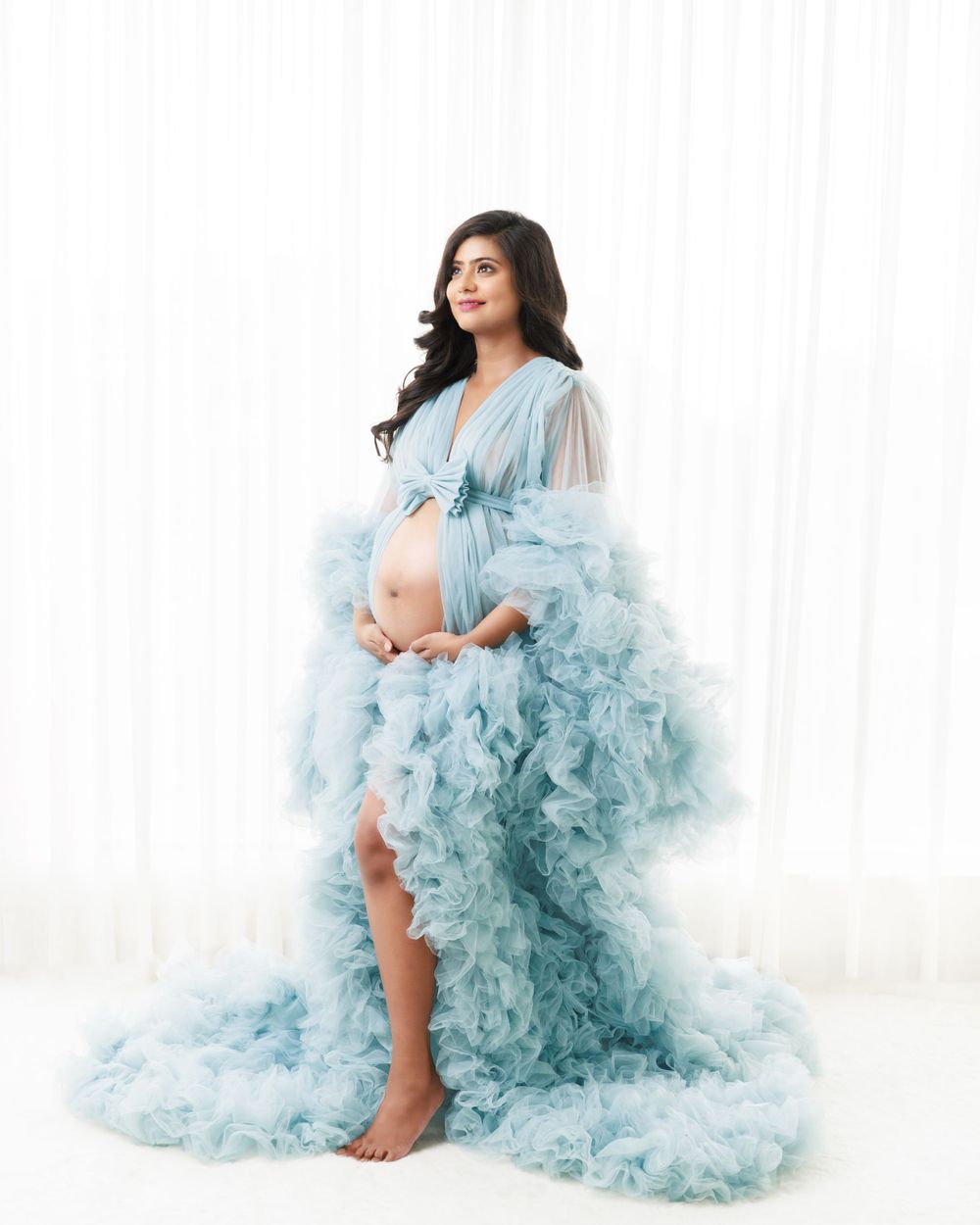 Dreamy maternity portrait in light blue ruffled tulle gown against white backdrop.