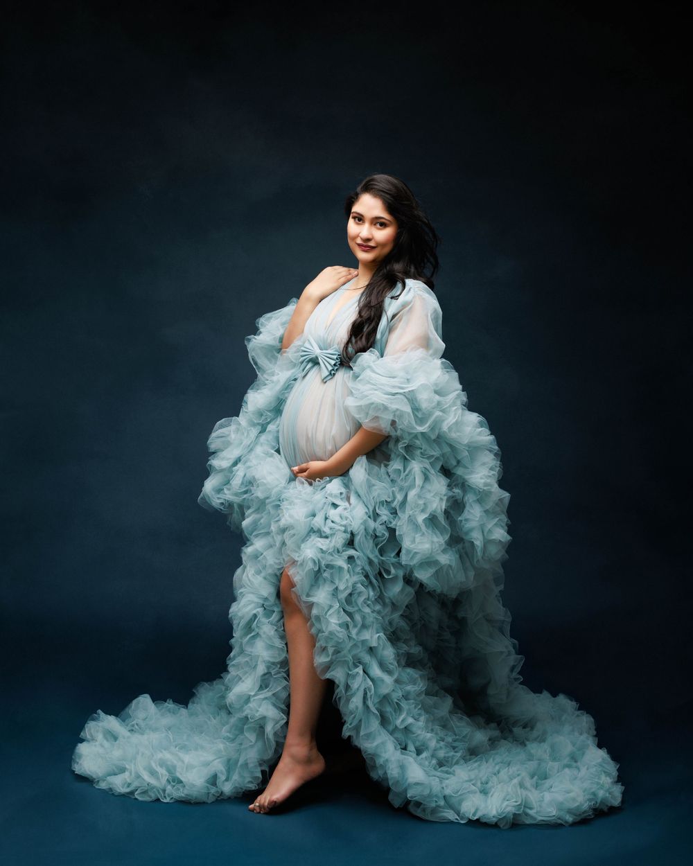 Ethereal maternity portrait in flowing light blue ruffle gown against dark backdrop.
