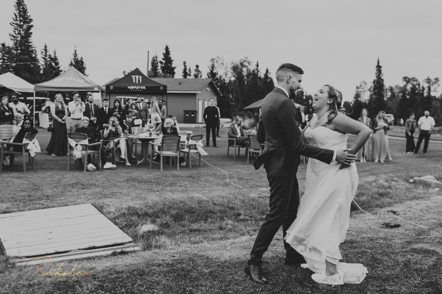Black and white photo of a couple dancing at an outdoor reception with guests in the background.