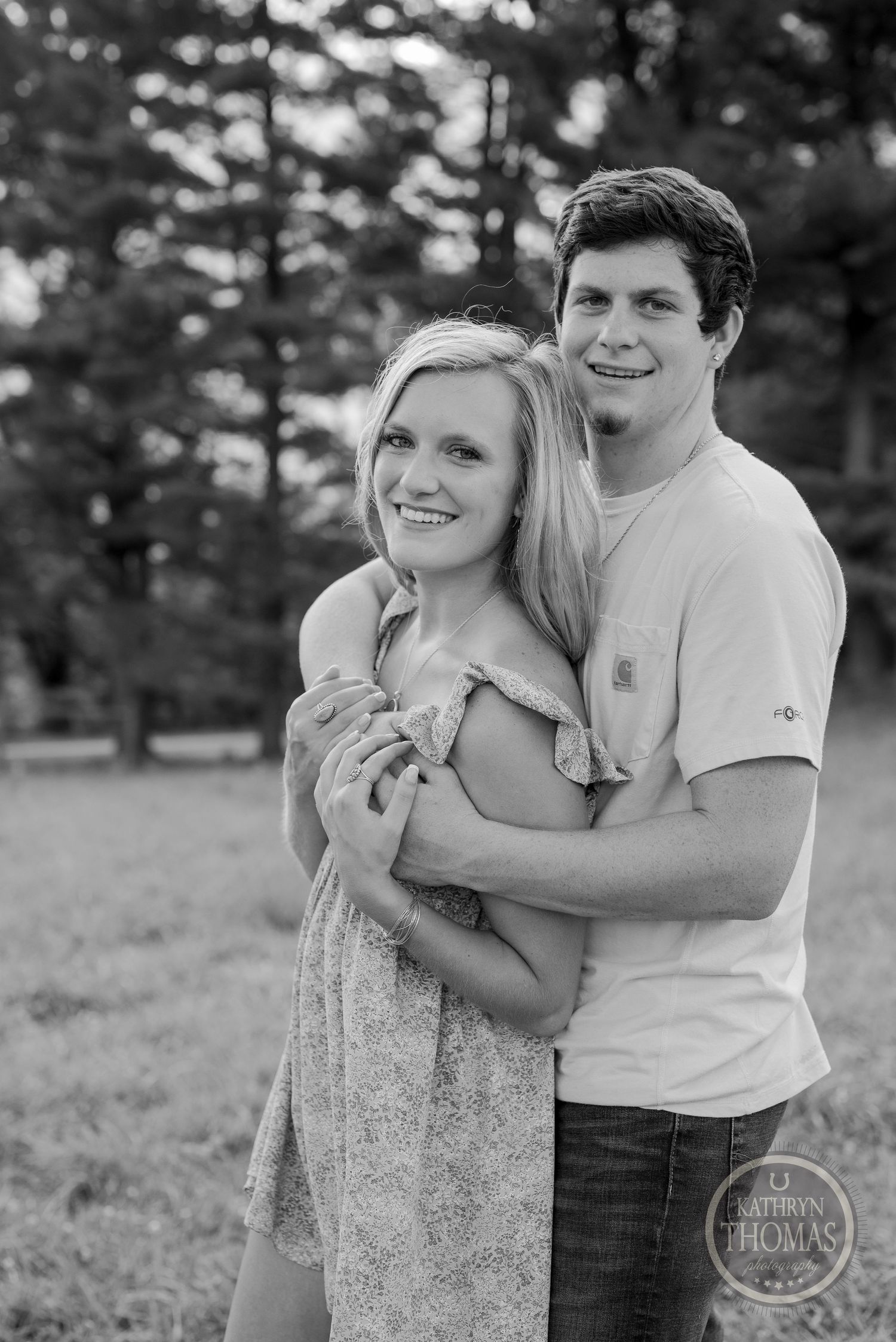 Black and white portrait of couple embracing outdoors with forest backdrop.