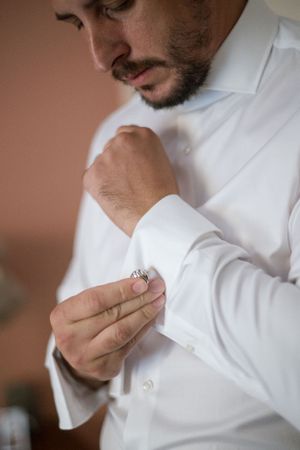 Groom in white shirt fastening cufflink during wedding preparation.
