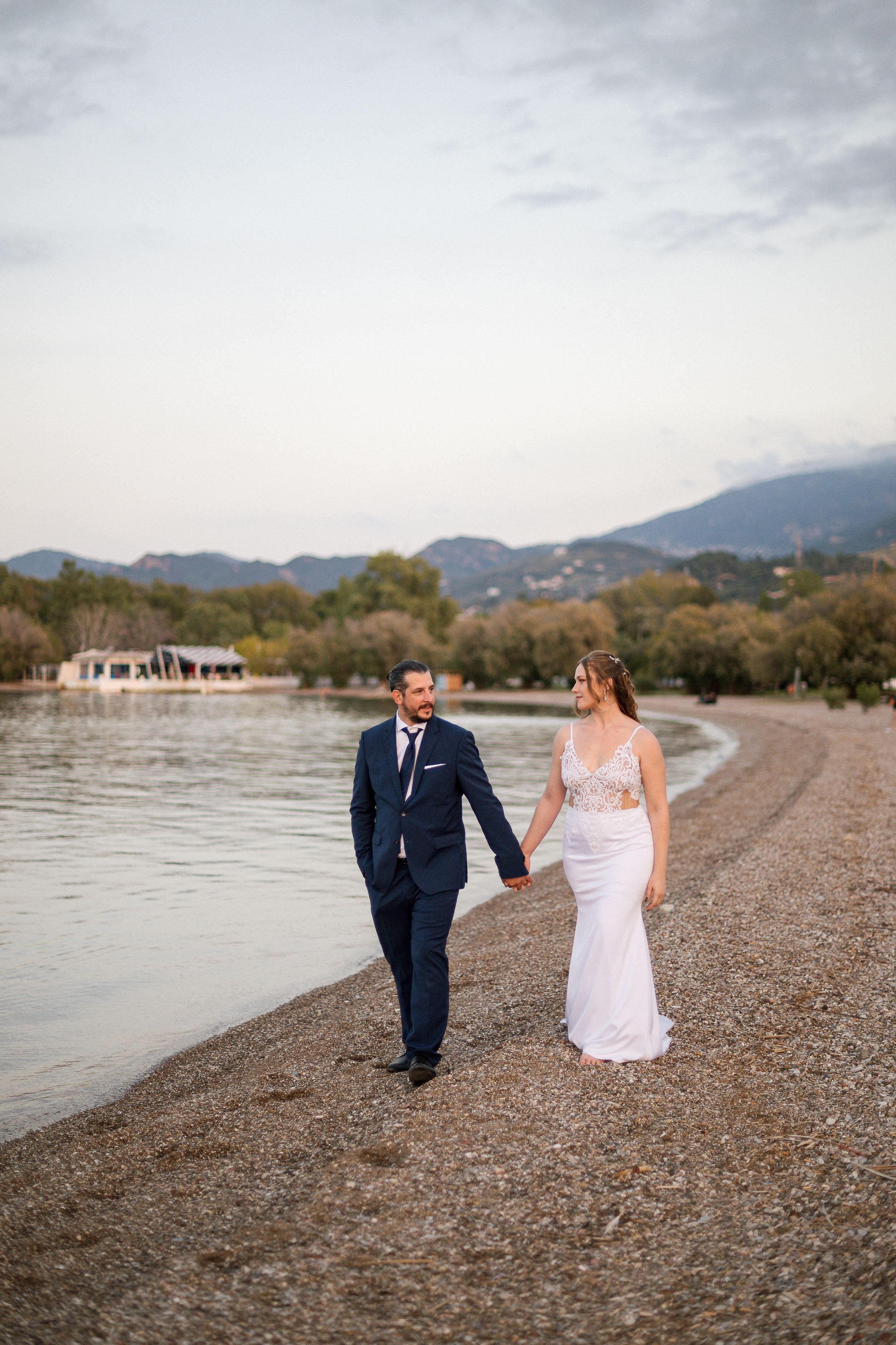 Couple walking hand in hand along lakeside beach with mountains in background.