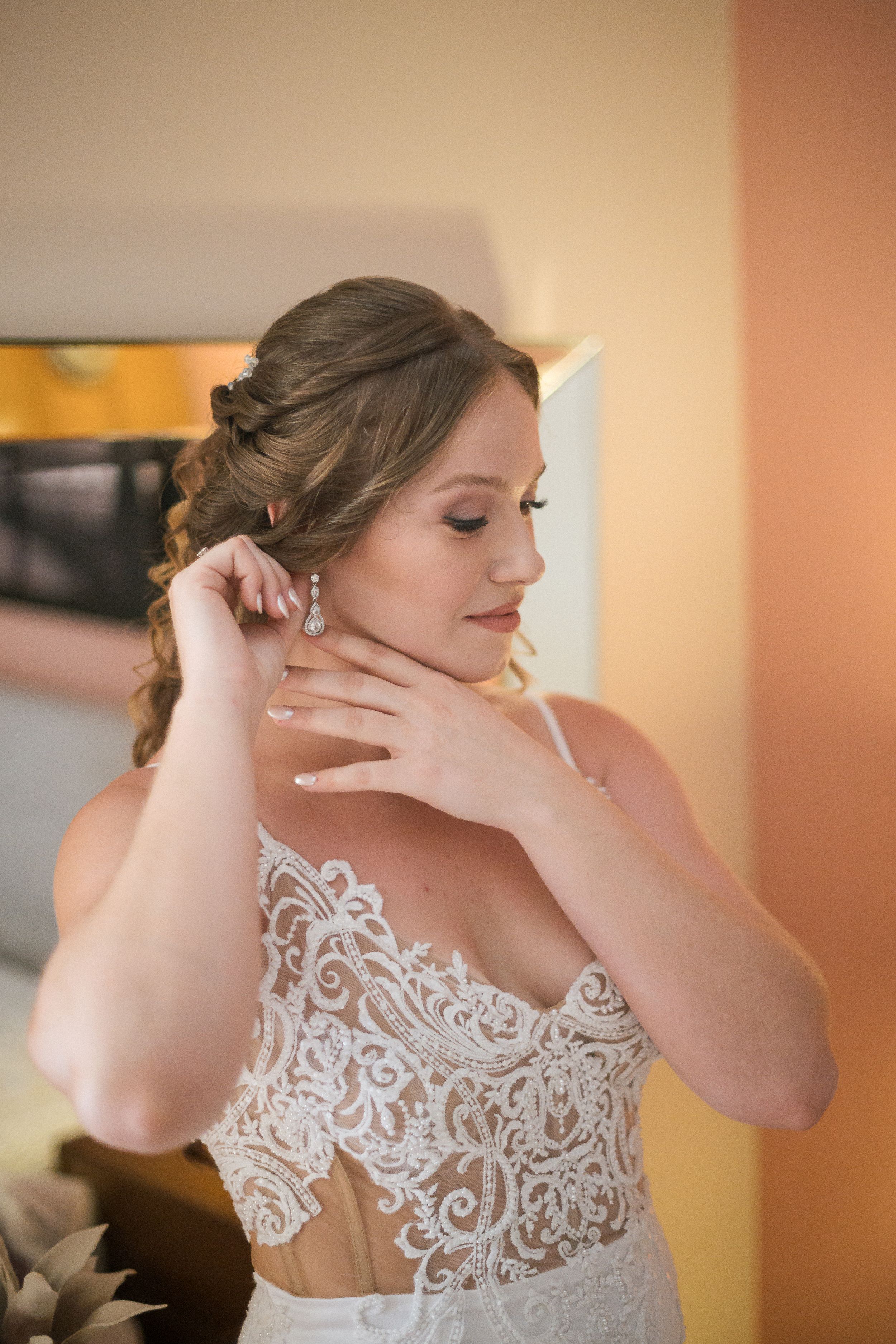 Bride adjusts her earring while getting ready in an elegant room with warm lighting.