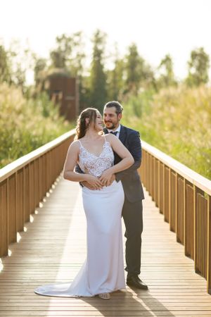 Wedding couple embraces on a wooden boardwalk surrounded by tall grass at sunset.