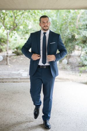 Professional groom in a navy suit and tie walking with confidence in an outdoor garden setting.