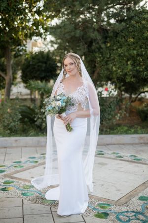 Bride in elegant white lace gown holding blue bouquet in garden setting.