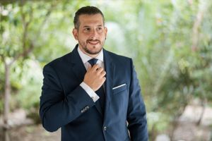 Elegant groom adjusts tie while standing in natural outdoor setting.