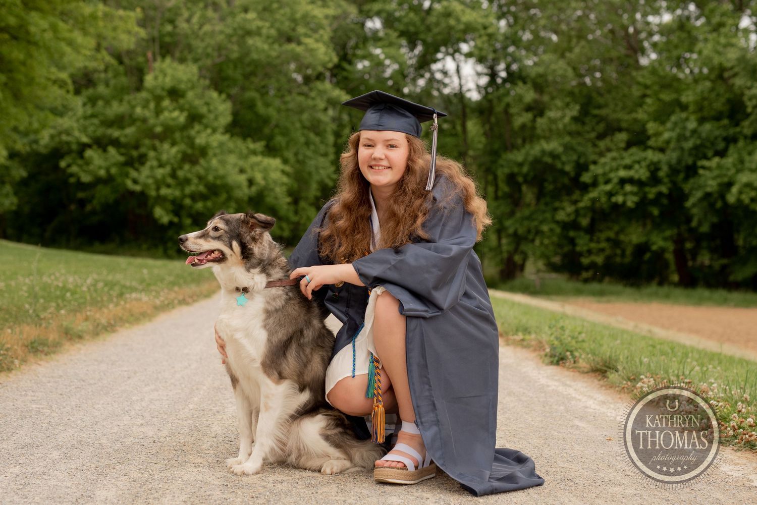 Graduate poses on dirt path with friendly husky mix dog while wearing cap and gown.