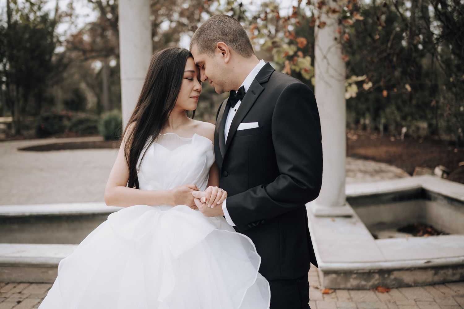 Bride in white gown and groom in tuxedo share a tender moment by stone columns.