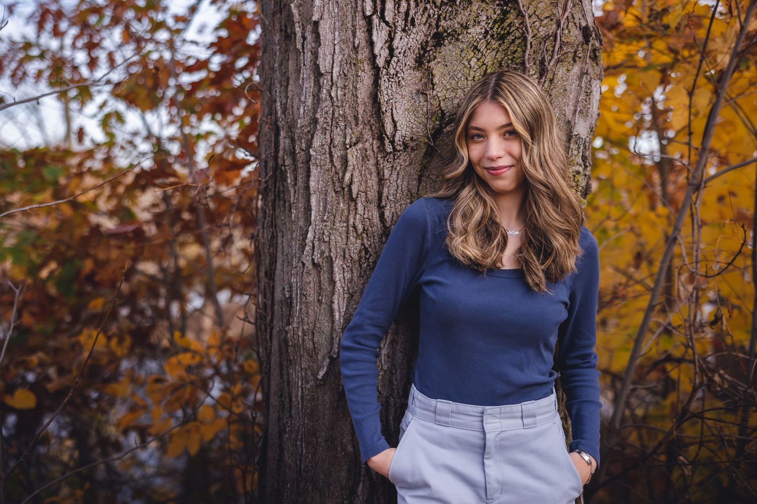 Woman in navy sweater leans against large tree trunk surrounded by autumn foliage.